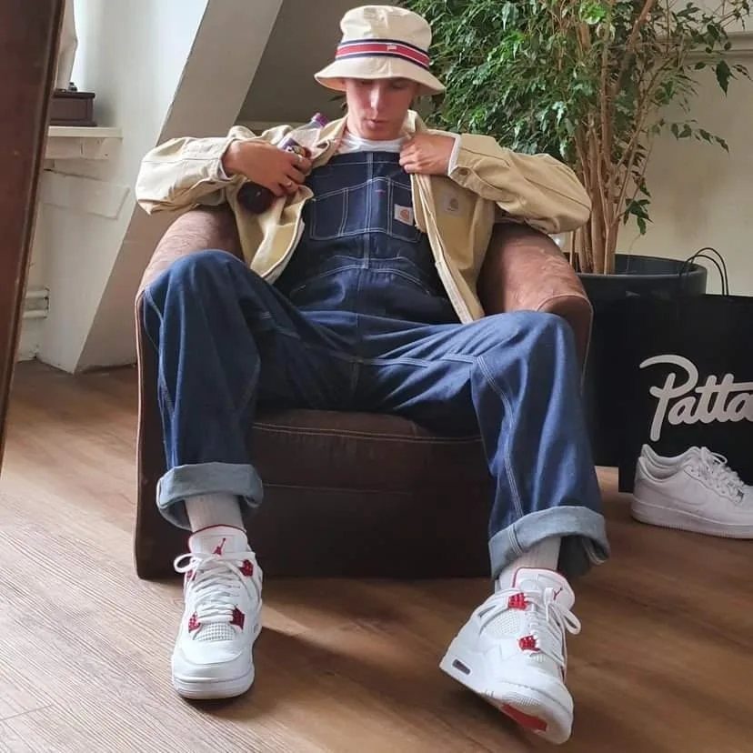 Person wearing baggy denim overalls, beige jacket, and bucket hat sitting in armchair.
