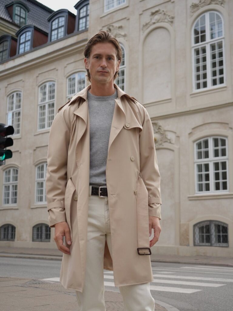 Man in a beige trench coat and white trousers standing before a white building.