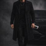 Man in a long black coat and trousers standing before a dark car