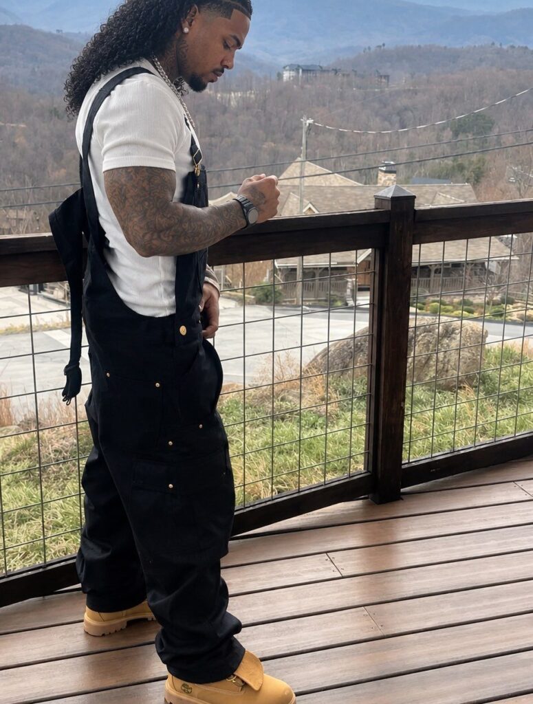 Man wearing black overalls, a white t-shirt, and tan boots on a deck.