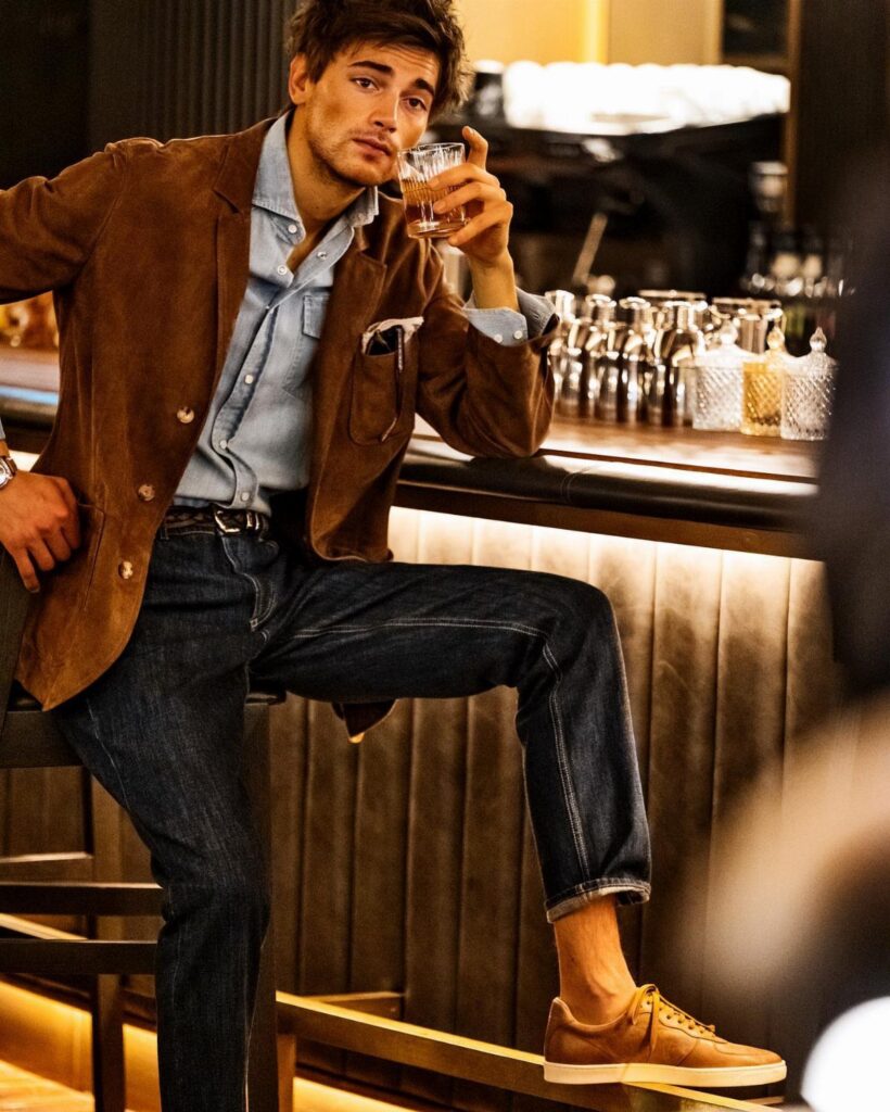 Man wearing a brown suede jacket and navy blue jeans sitting at a bar.