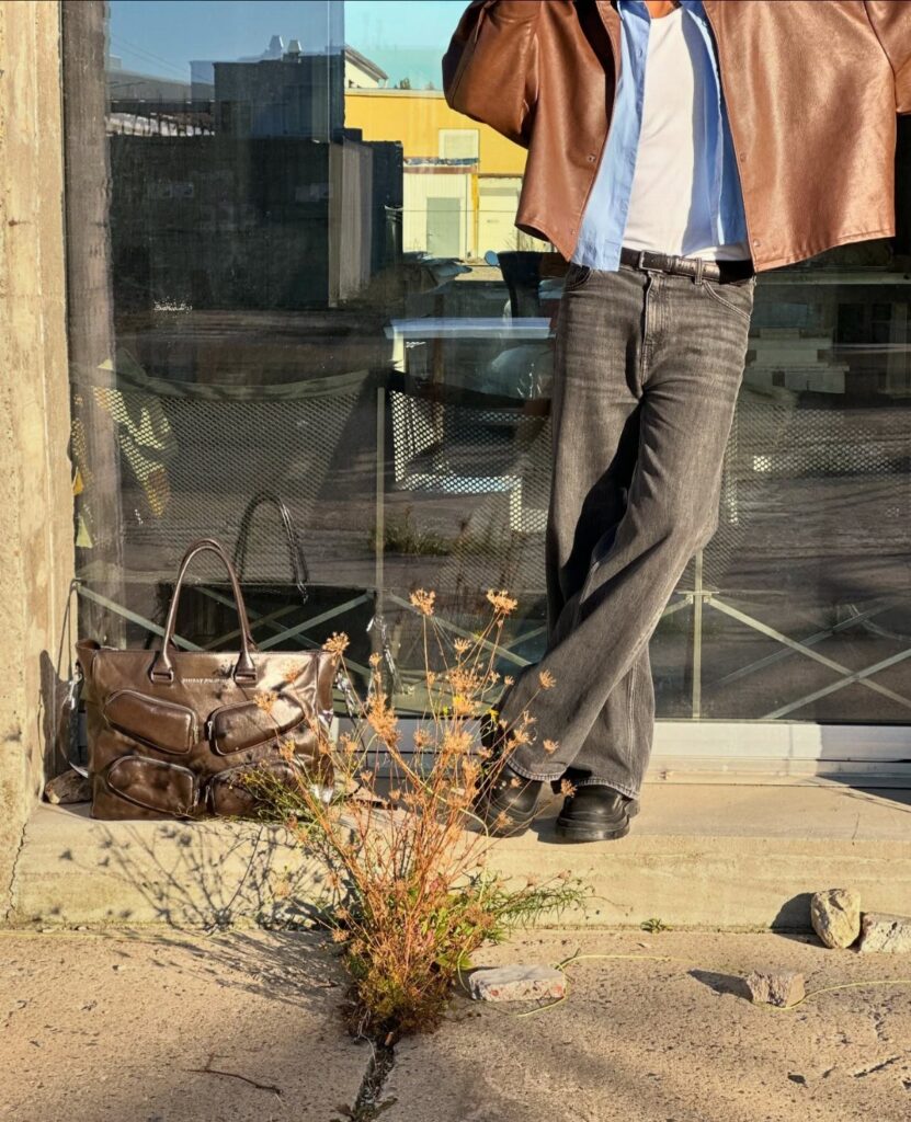 Person wearing a brown leather jacket, blue shirt, and grey jeans outdoors.
