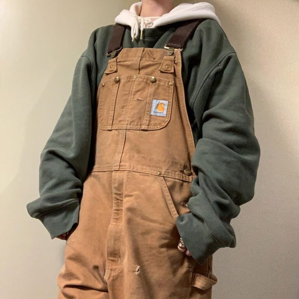 Person wearing brown canvas overalls over a green sweatshirt and white hoodie.