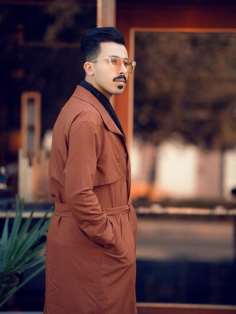 Man wearing a brown trench coat and yellow glasses posing outdoors.