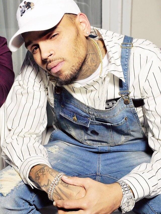 Chris Brown wearing blue denim overalls, a striped shirt, and a white cap.
