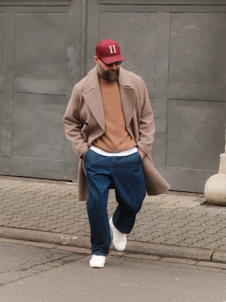 Man wearing a beige overcoat and blue jeans walking on a city street.