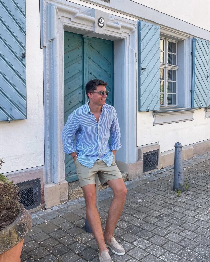 Man in striped long-sleeve shirt and beige shorts leaning against a white building.
