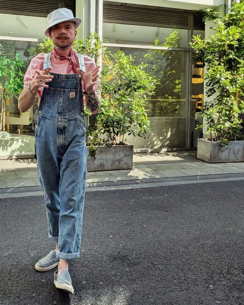 Man wearing blue denim overalls, pink shirt, and white bucket hat on a street