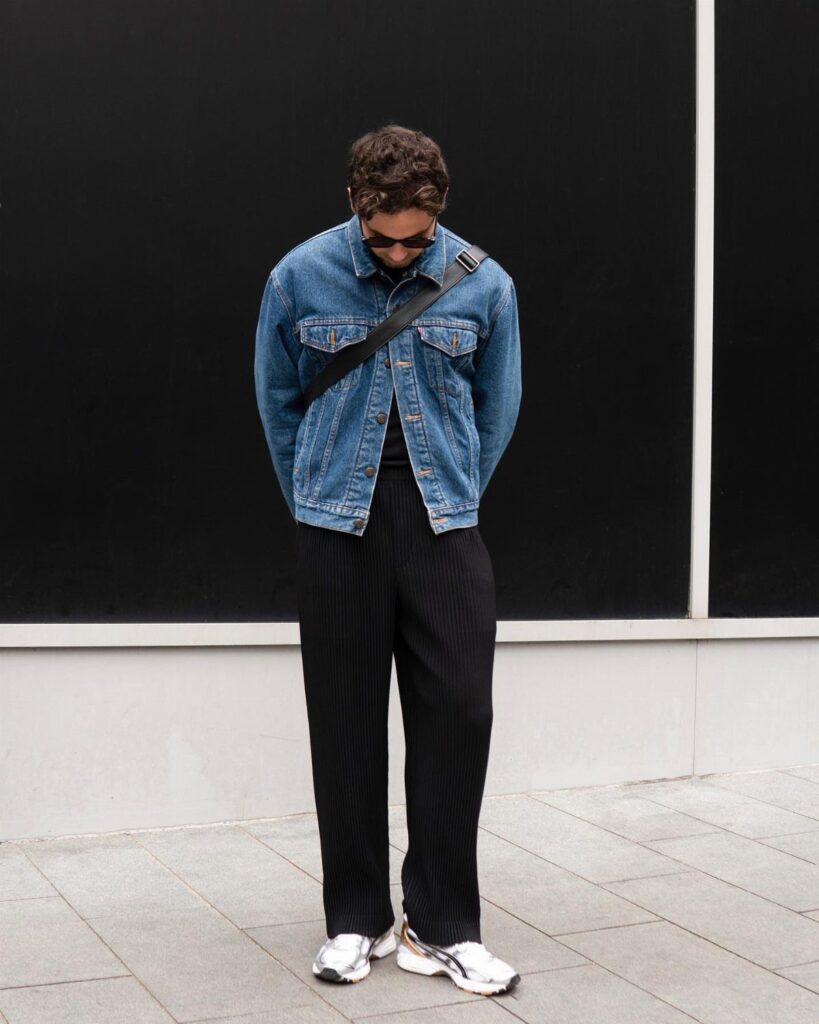 Person wearing a blue denim jacket and black wide-leg trousers against a black wall.