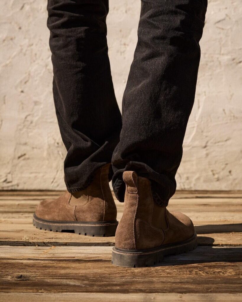Brown suede ankle boots with thick rubber soles and black trousers on wood.
