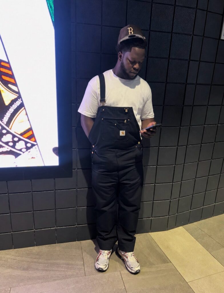 Man wearing black overalls over a white t-shirt and white sneakers.