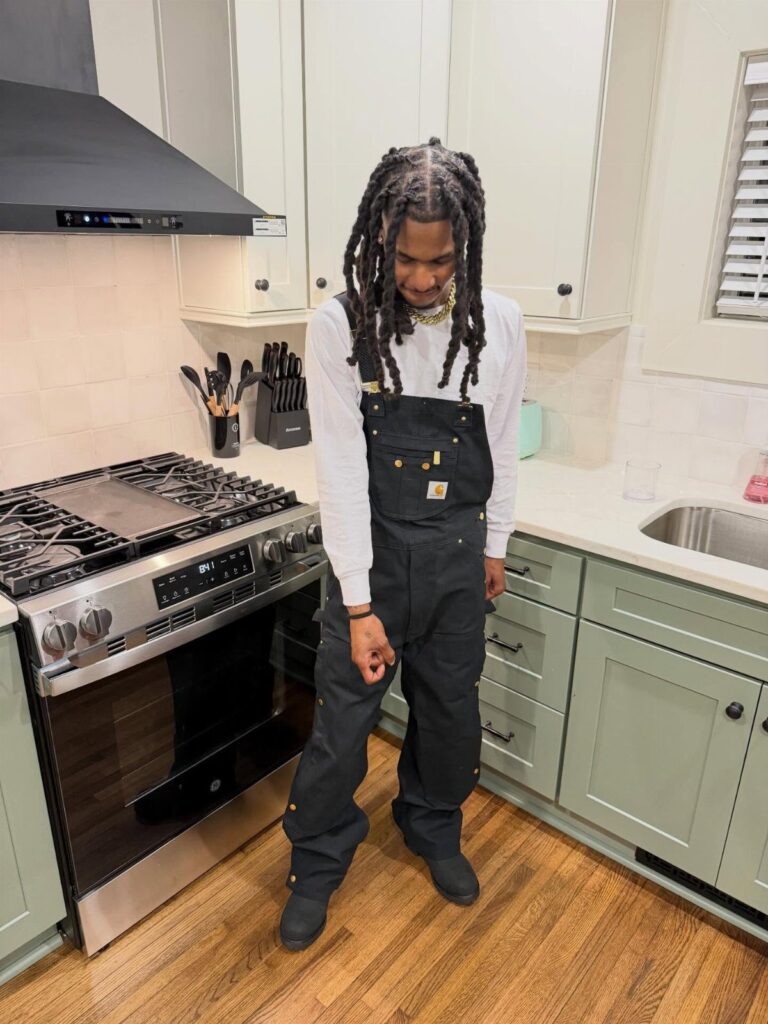 Man wearing black overalls over a white shirt standing in a modern kitchen.