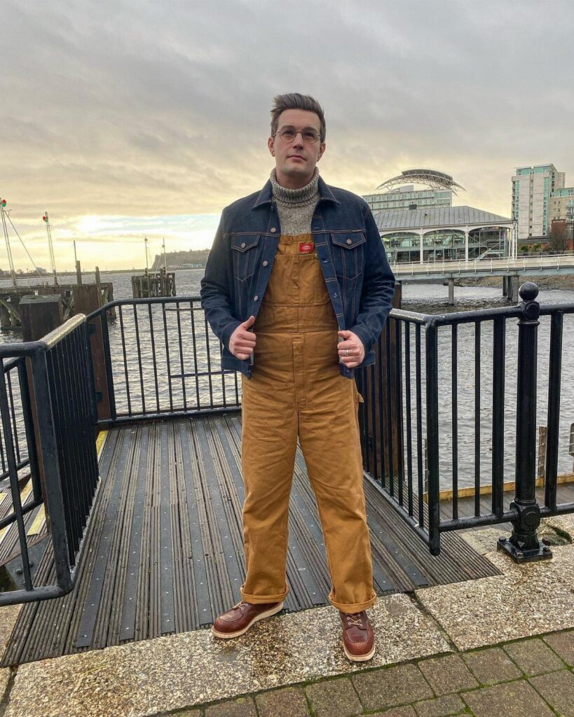 Man wearing brown dungarees over a grey chunky knit turtleneck and denim jacket.