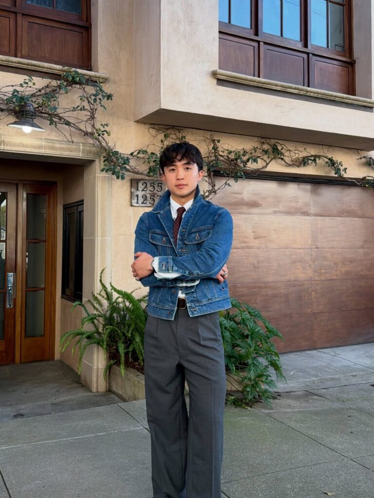 Man wearing a blue denim jacket and grey tailored trousers standing outdoors.