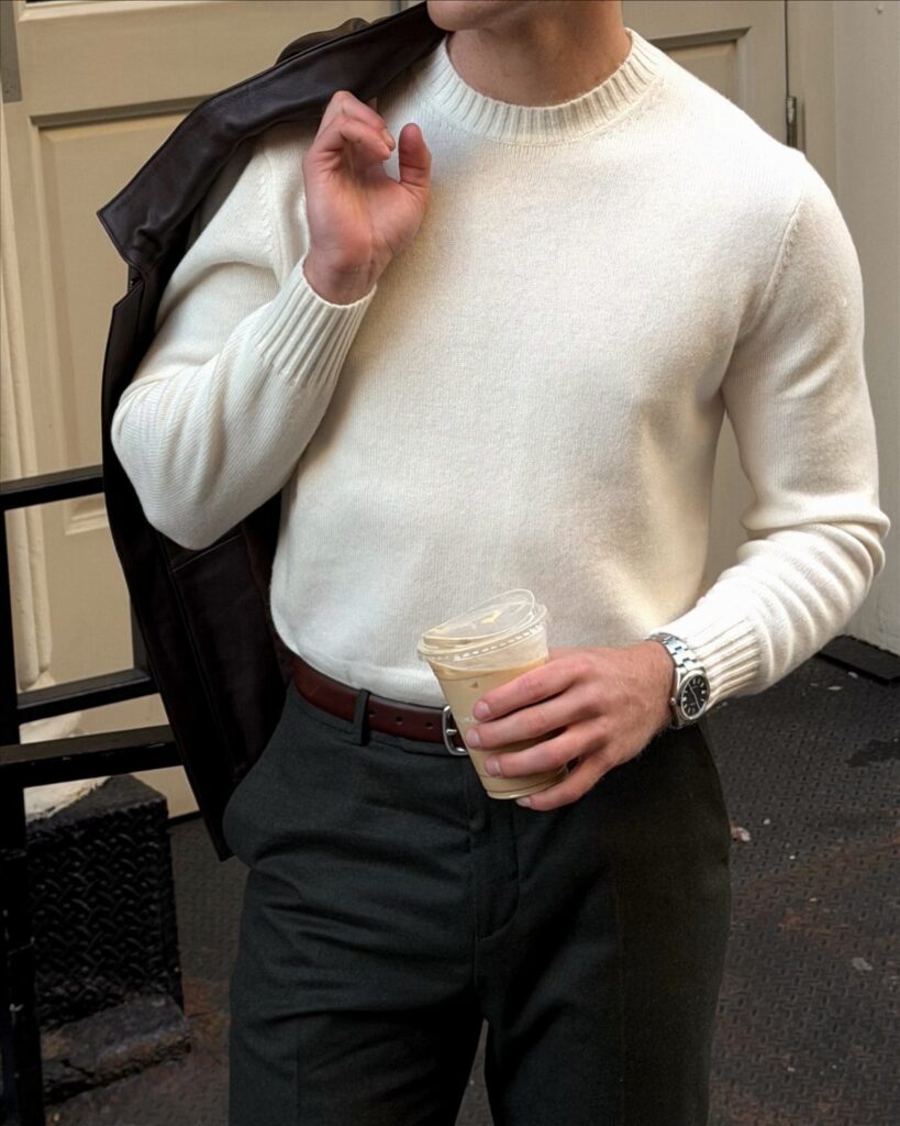 Man in white sweater and grey trousers holding a brown leather jacket.
