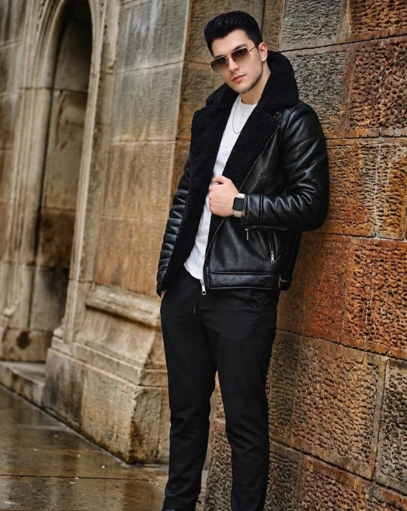 Man in black leather shearling jacket and black trousers leaning against stone wall