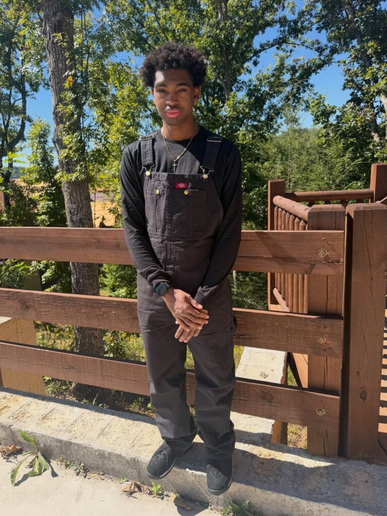 Young man wearing black overalls and a black shirt standing by a wooden fence