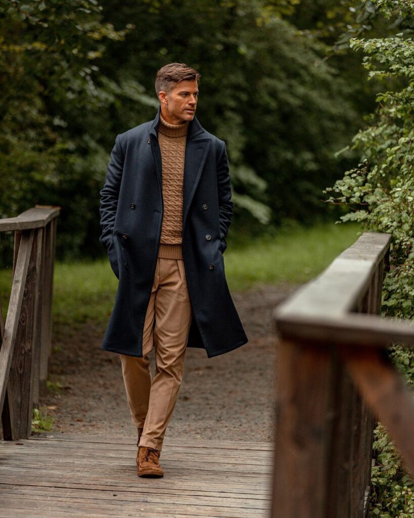Man in navy blue wool coat and tan sweater walking on wooden bridge