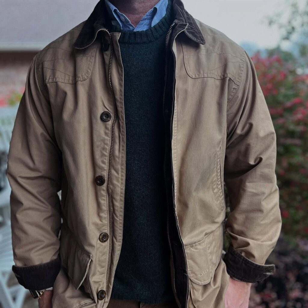 Tan field coat with brown collar worn over a green sweater and blue shirt.