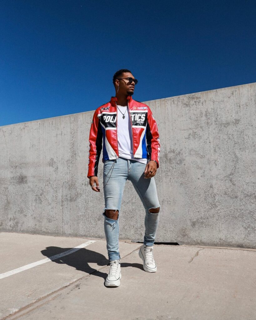 Man wearing a red, white, and blue racer jacket with ripped light-blue jeans.