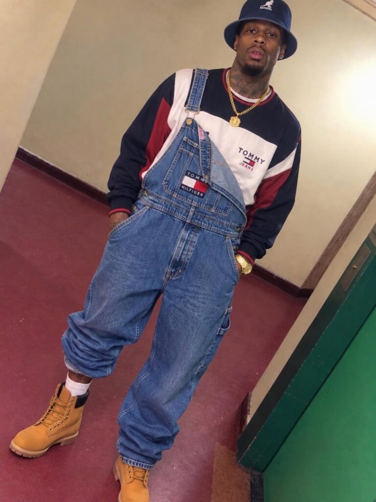 Man wearing blue denim overalls, a tri-color sweatshirt, bucket hat, and tan boots.