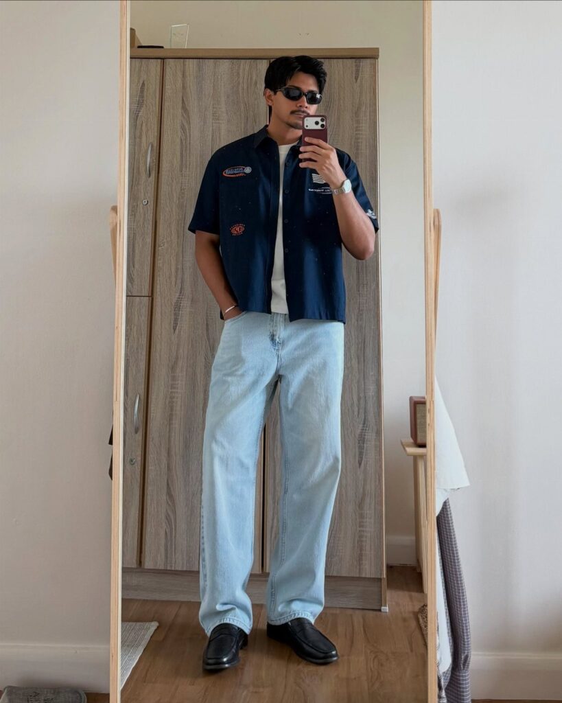 Man in blue short-sleeved shirt and light blue jeans taking a mirror selfie.