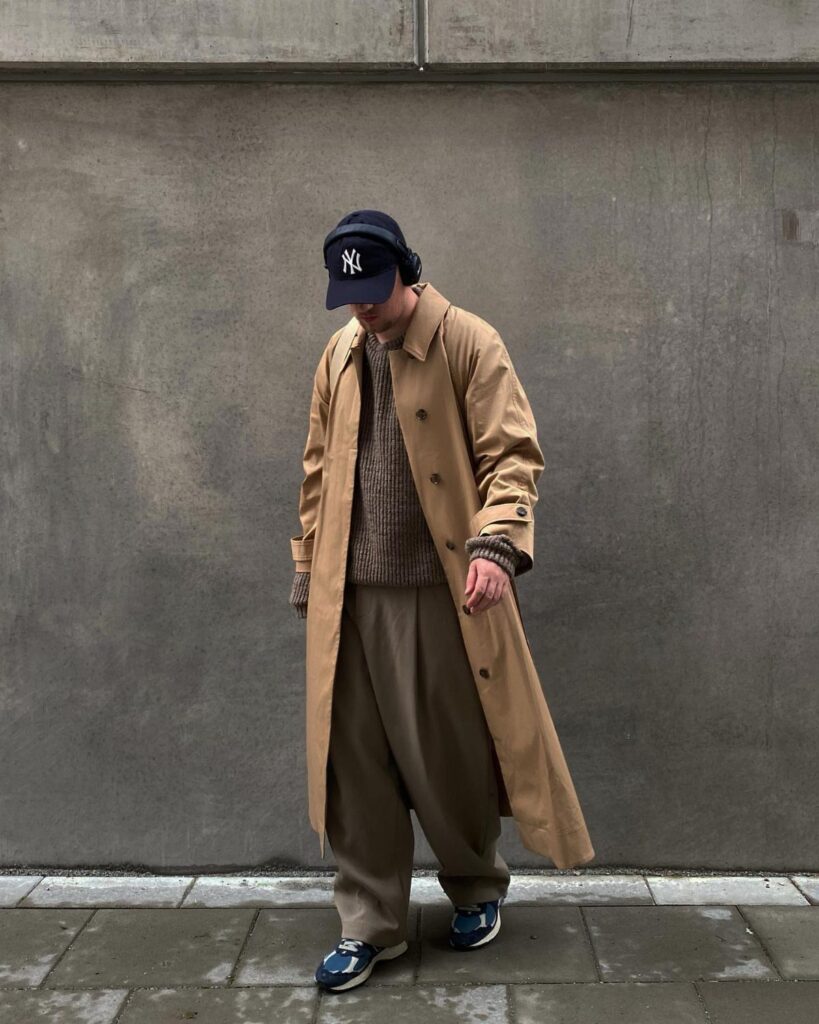 Person wearing a tan trench coat, brown sweater, and khaki trousers against concrete.