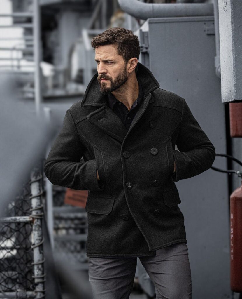 Man wearing a dark grey double-breasted wool peacoat on a ship deck.