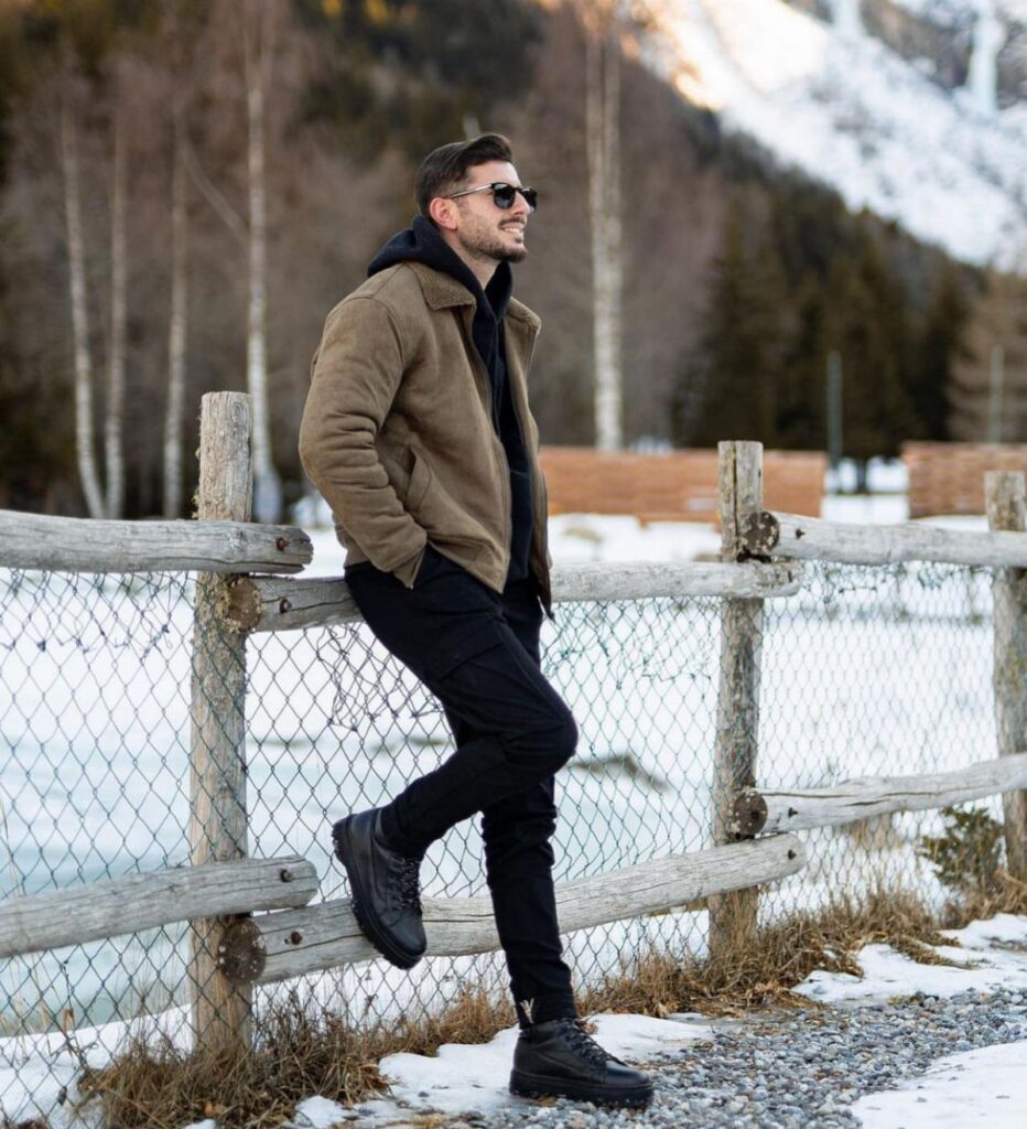 Man in brown winter jacket and black pants leaning on a wooden fence