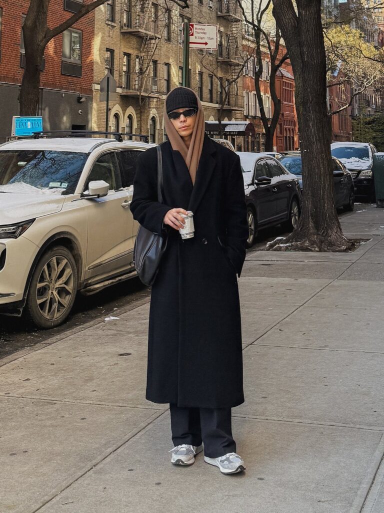 Person wearing a long black wool coat, brown scarf, and beanie on a city street
